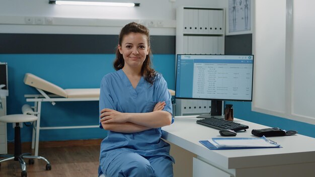 Retrato de asistente médico de pie con los brazos cruzados en el gabinete para una visita de revisión médica. Enfermera que trabaja con computadoras y documentos en el consultorio médico para el sistema de salud.