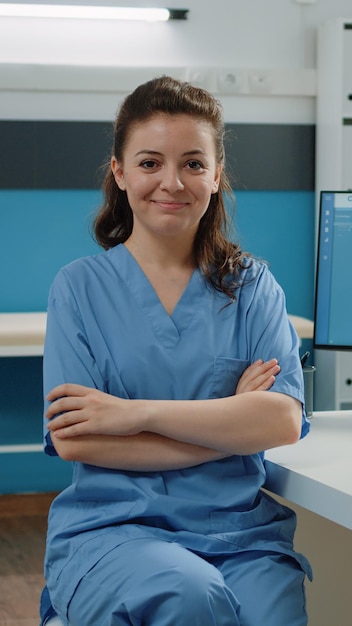 Retrato de asistente médico de pie con los brazos cruzados en el gabinete para una visita de revisión médica. Enfermera que trabaja con computadoras y documentos en el consultorio médico para el sistema de salud.