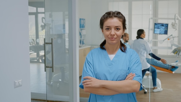 Retrato de asistente de odontología mirando a la cámara de pie en la clínica de estomatología. Mujer caucásica con ocupación de enfermera trabajando en consultorio dental vistiendo uniforme médico para el cuidado de los dientes