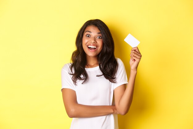 Retrato de asombrada niña afroamericana levantando tarjeta de crédito y sonriendo emocionado yendo de compras