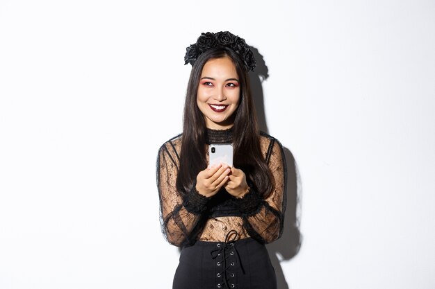 Retrato de astuta hermosa mujer asiática, bruja en vestido de encaje gótico con teléfono móvil, sonriendo y mirando a la esquina superior izquierda, de pie sobre fondo blanco.