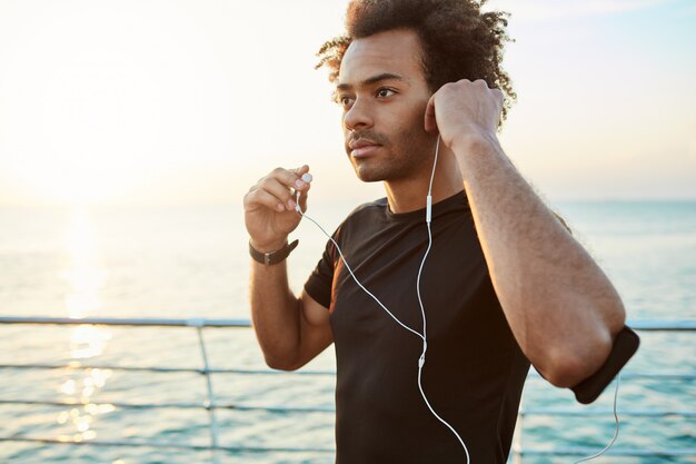 Retrato de atleta afroamericano con auriculares blancos, preparándose para trotar. Corriendo al amanecer detrás del mar. Concepto de estilo de vida saludable