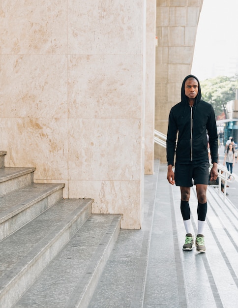 Retrato de un atleta masculino joven africano en sudadera con capucha negra que se coloca cerca de los pasos