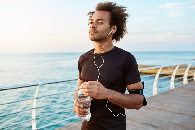 Retrato de un atleta masculino de piel oscura concentrada y mediadora con cabello tupido sosteniendo una botella de agua mineral en sus manos.