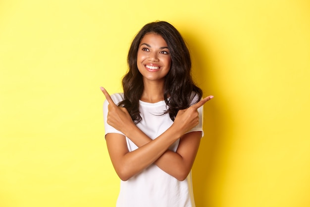 Retrato de atractiva chica afroamericana en camiseta blanca sonriendo y mirando hacia la izquierda apuntando