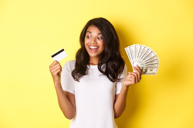 Retrato de atractiva chica afroamericana sosteniendo dinero y mirando la tarjeta de crédito permanente