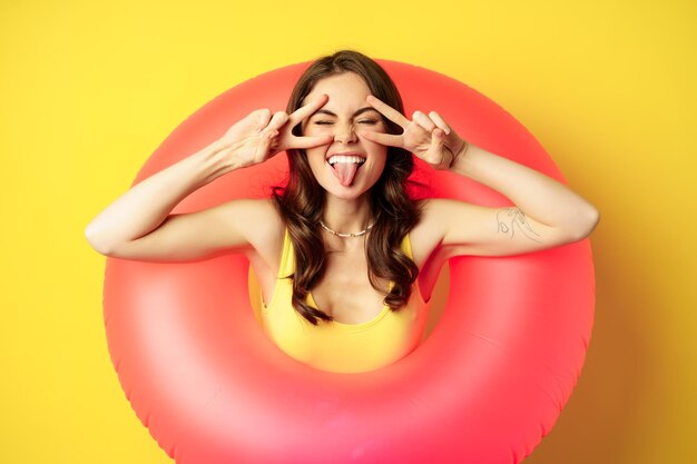 Retrato de una atractiva chica moderna dentro de un anillo de natación de playa rosa de moda, mostrando el signo de la paz y sonriendo feliz, de pie sobre un fondo amarillo