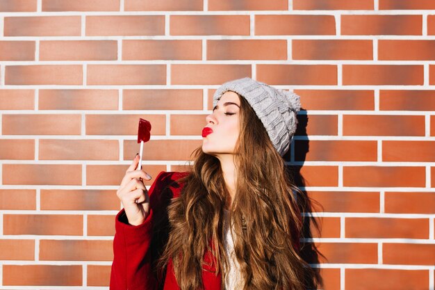 Retrato atractiva chica morena en la pared exterior. Lleva gorro de punto, abrigo rojo. Sostiene los labios rojo caramelo, mantiene los ojos cerrados y envía besos.