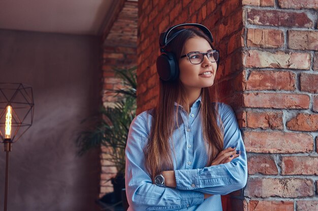 Retrato de una atractiva y encantadora morena con gafas y pantalones azul cruzado de brazos y escuchando música con auriculares apoyados en una pared de ladrillos en una habitación con diseño de loft.