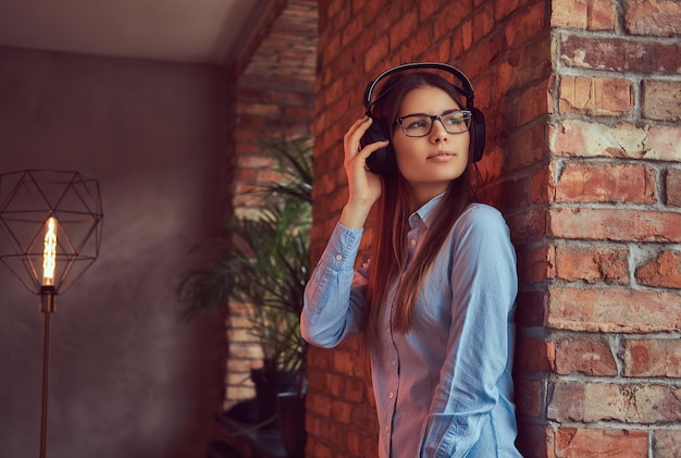 Retrato de una atractiva y encantadora morena con gafas y pantalones azul escuchando música con auriculares apoyados en una pared de ladrillos en una habitación con diseño de loft.