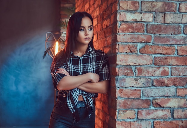 Retrato de una atractiva hipster con camisa de franela apoyada en la pared en una habitación con interior de loft.