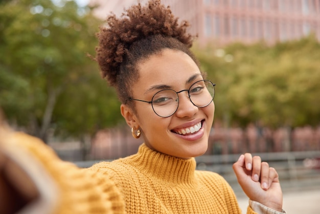 Retrato de una atractiva joven sonriente de cabello rizado con anteojos redondos ópticos posa para selfies al aire libre camina en un entorno urbano durante las fotografías de tiempo libre ella misma tiene un día libre o un fin de semana