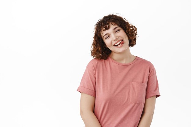 Retrato de una atractiva joven sonriente, con la cabeza inclinada linda y mostrando la lengua graciosa, mostrando una sonrisa positiva y feliz, de pie contra un fondo blanco.