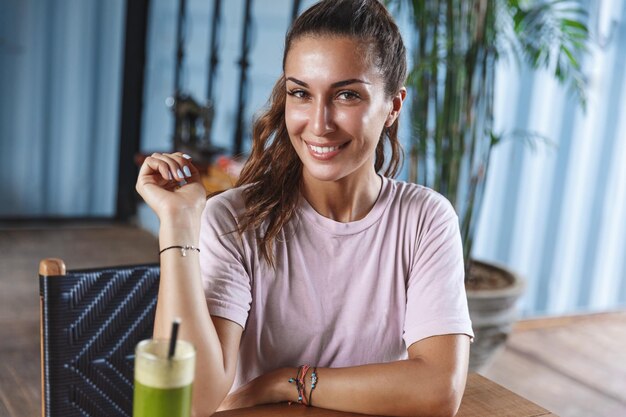 Retrato de una atractiva joven turista bronceada y coqueta relajándose en la isla de Bali sentada en un café de mesa de madera bebiendo batido y disfrutando de la vista del soleado resort Pradise sonriendo feliz a la cámara