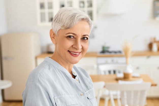 Retrato de atractiva jubilada caucásica senior con estilo con peinado corto pixie pasar el día en casa, de pie en la sala de estar con un elegante vestido azul, sonriendo felizmente