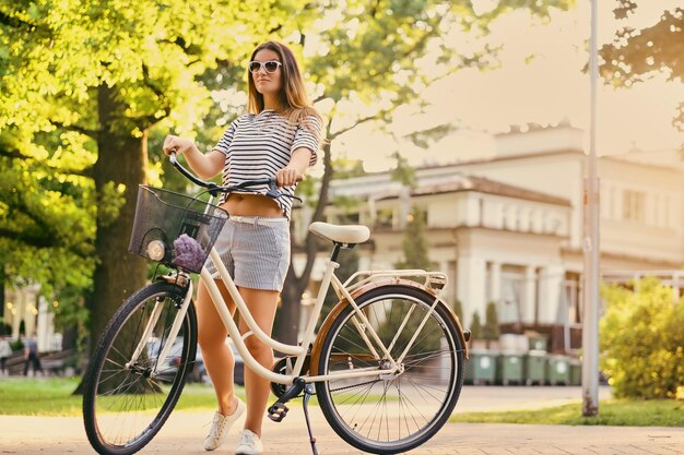 Retrato de una atractiva morena con una bicicleta en el parque de la ciudad.