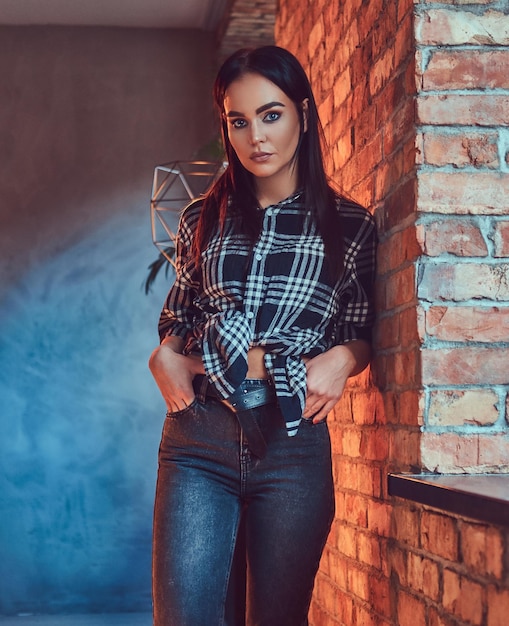 Retrato de una atractiva morena vestida con una camisa de franela y jeans apoyados en la pared en una habitación con interior de loft.