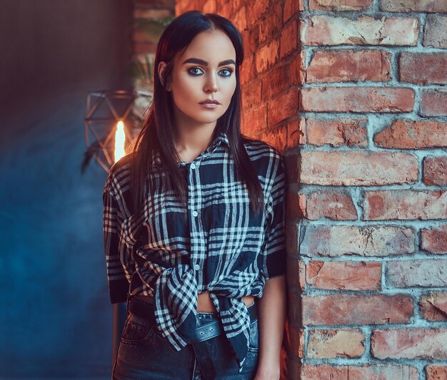 Retrato de una atractiva morena vestida con una camisa de franela y jeans apoyados en la pared en una habitación con interior de loft.