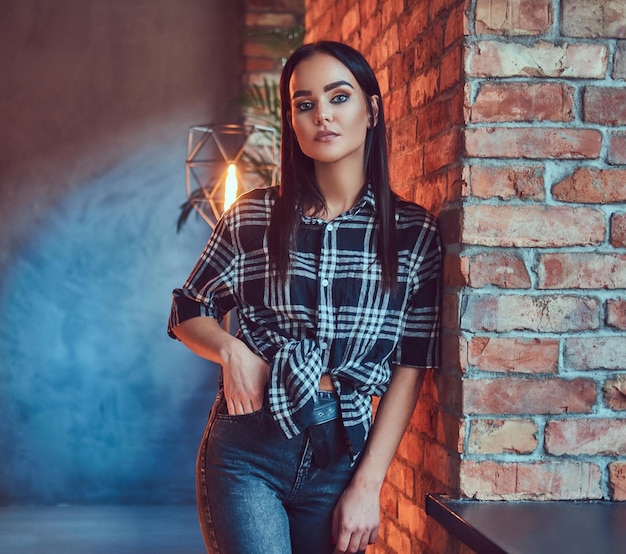 Retrato de una atractiva morena vestida con una camisa de franela y jeans apoyados en la pared en una habitación con interior de loft.