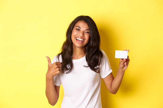 Retrato de atractiva mujer afroamericana en camiseta blanca mostrando thumbsup y tarjeta de crédito