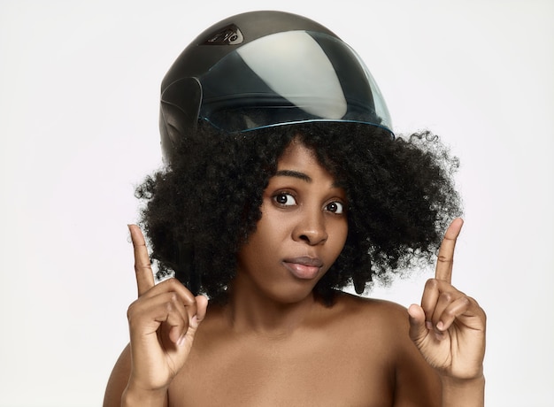 Retrato de atractiva mujer afroamericana sorprendida en casco de moto sobre fondo blanco de estudio. Concepto de belleza y protección de la piel.