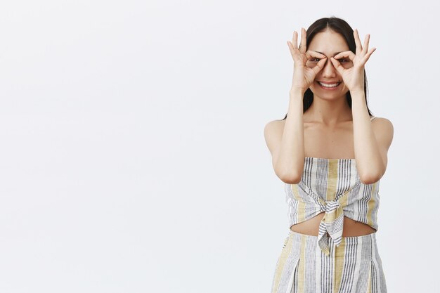Retrato de atractiva mujer asiática alegre con cabello largo y oscuro en ropa de verano a juego de moda, haciendo gestos de ojos con los dedos y mirando como en gafas