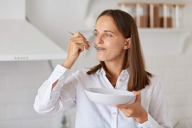 Retrato de atractiva mujer hambrienta con cabello oscuro con camisa blanca, mirando a otro lado, posando en la cocina, desayunando, sosteniendo el plato y la cuchara en las manos.