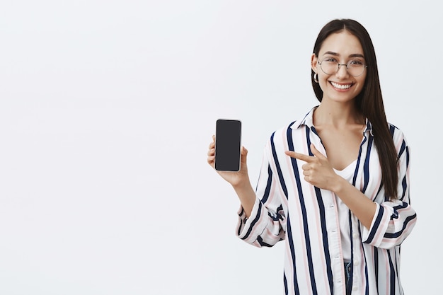 Retrato de atractiva mujer de moda complacida con gafas y blusa, mostrando la pantalla en el teléfono y apuntando al dispositivo con el dedo índice, sonriendo ampliamente