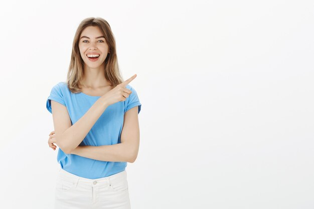 Retrato de atractiva mujer rubia alegre sonriendo divertido y apuntando hacia la esquina superior derecha, mostrando el camino