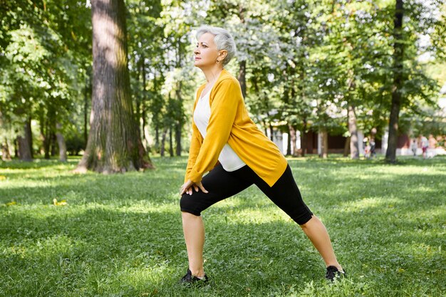 Retrato de atractiva mujer sana de sesenta años de pie sobre una pierna y estiramiento en pose de pilates. Mujer mayor de pelo gris en ropa deportiva haciendo estocadas laterales sobre el césped en el parque de la ciudad en un día soleado