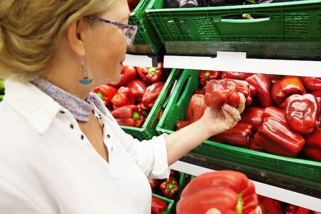 Retrato de una atractiva pensionista que compra frutas y verduras en el departamento de productos de la tienda de comestibles o supermercado, recogiendo grandes pimientos rojos para la cena familiar, eligiendo los mejores