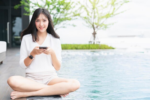 Retrato de un atractivo asiático de pelo largo y negro, vestido casual, sonrisa, uso de la mano, teléfono inteligente, sentarse, relajarse cerca del jardín de la piscina con el concepto de ideas de comunicación y tecnología de fondo de la ciudad