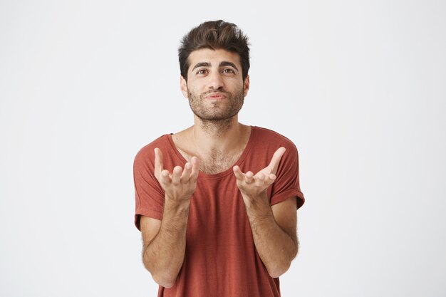 Retrato de atractivo estudiante barbudo en camiseta roja estirando sus manos. Hombre emocional con peinado elegante que no entiende lo que se quiere de él