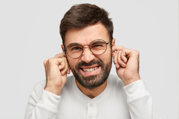 Retrato de atractivo hombre sin afeitar tapona los oídos mientras escucha un sonido molesto, frunce el ceño, aprieta los dientes