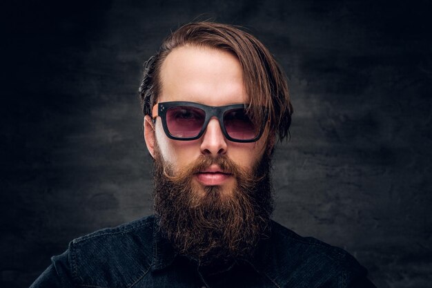 Retrato de un atractivo hombre barbudo con gafas de sol sobre un fondo oscuro.
