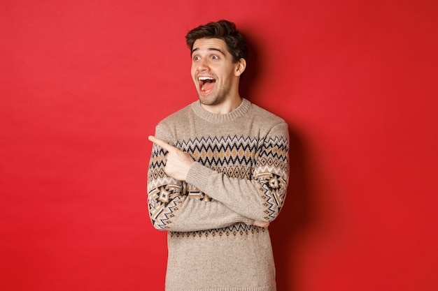 Retrato de atractivo hombre caucásico celebrando año nuevo, vacaciones de invierno, señalando con el dedo y mirando a la izquierda con expresión de asombro, mostrando publicidad navideña, de pie sobre fondo rojo.