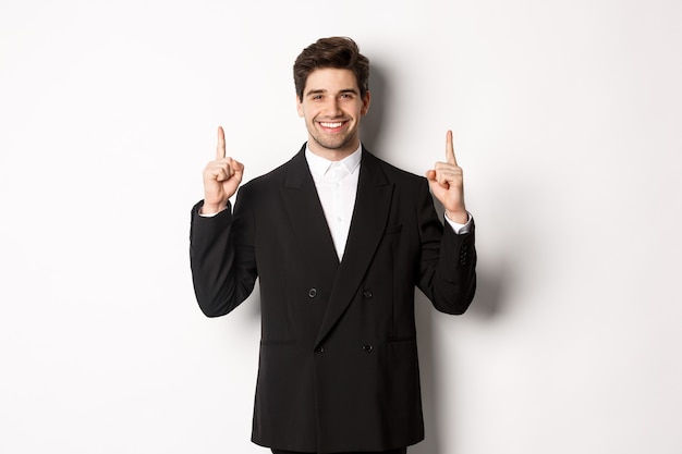 Retrato de atractivo hombre caucásico en elegante traje negro, apuntando con el dedo hacia arriba y sonriendo, mostrando publicidad navideña, de pie sobre fondo blanco.