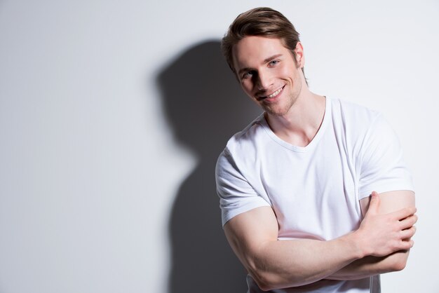 Retrato de atractivo hombre sonriente en camiseta blanca plantea sobre la pared con sombras de contraste.