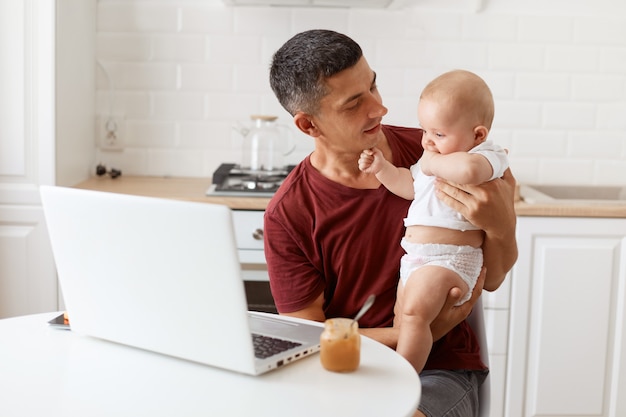 Retrato de atractivo independiente hombre morena vistiendo camiseta granate de estilo casual, trabajando y cuidando a su pequeña hija, mirando al bebé con amor.