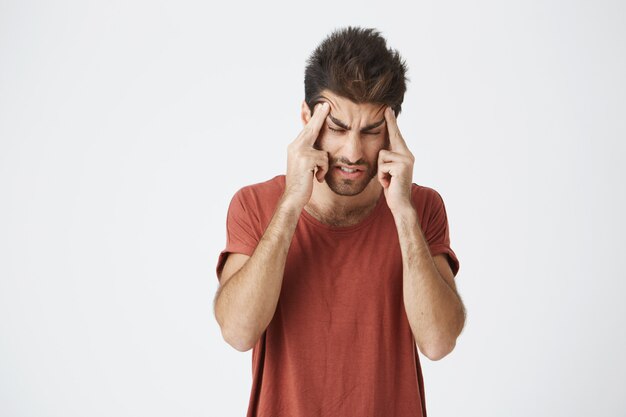 Retrato de atractivo joven español vistiendo una camiseta roja con la cara estresante y fruncida, apretando la cabeza con las manos teniendo dolor de cabeza después de dormir solo durante unas pocas horas.