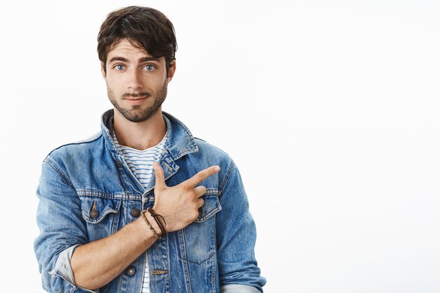 Retrato de atractivo joven hispano con barba y ojos azules apuntando a la esquina superior derecha sonriendo haciendo una pregunta, consultando a un amigo si se enteró de un nuevo café cercano, posando sobre una pared gris