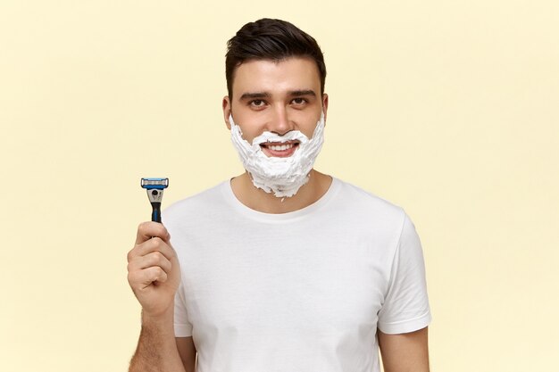 Retrato de atractivo joven de pelo oscuro en camiseta casual posando con crema de afeitar en el rostro, sosteniendo navaja desechable