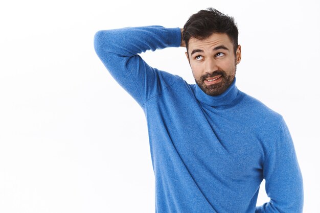 Retrato de un atractivo novio barbudo desorientado e indeciso con cuello alto, rascarse la cabeza y mirar hacia arriba, evitar el contacto visual, confesar que olvidó hacer la tarea, de pie en una pared blanca sin idea