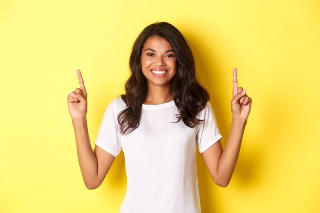 Retrato de atractivo y seguro modelo femenino afroamericano vistiendo camiseta blanca apuntando