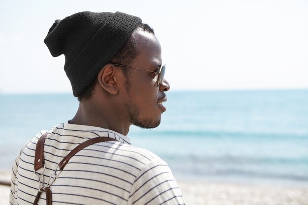 Retrato desde atrás de feliz viajero afroamericano de pie en la playa con mochila sobre sus hombros, con sombrero y gafas de sol, mirando al mar con esperanza, admiración, soñando con un futuro feliz