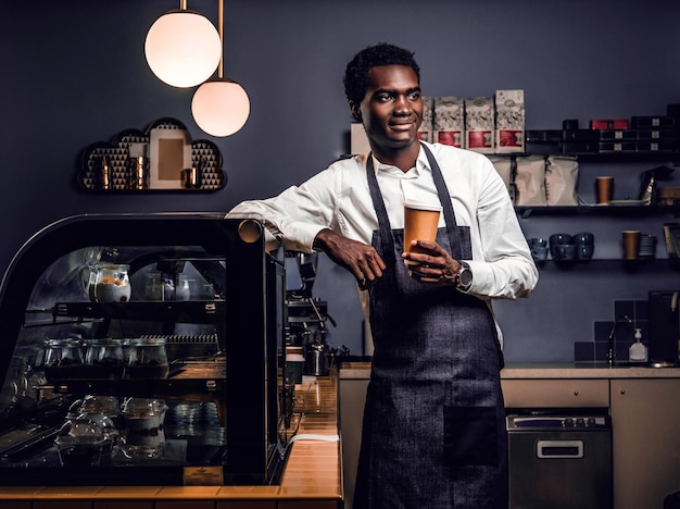 Foto gratuita retrato de un barista africano sosteniendo una taza de café mientras se apoya en un mostrador en una cafetería