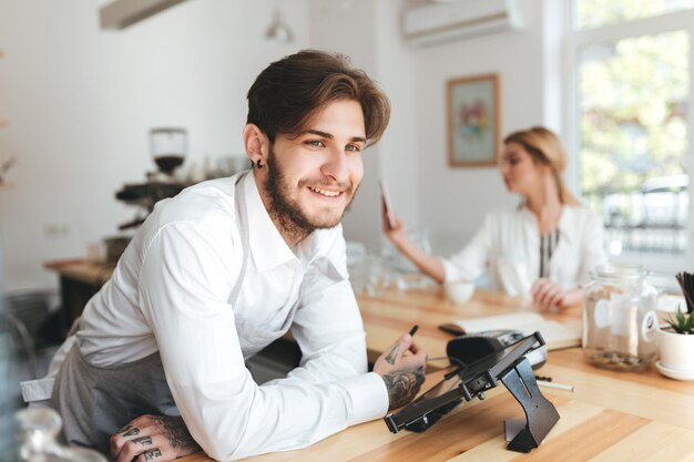 Retrato de barista sonriente en delantal y camisa blanca en el lugar de trabajo en la cafetería. Barista mirando felizmente a un lado de pie en el mostrador mientras una chica bonita en el fondo usa su teléfono móvil en el café