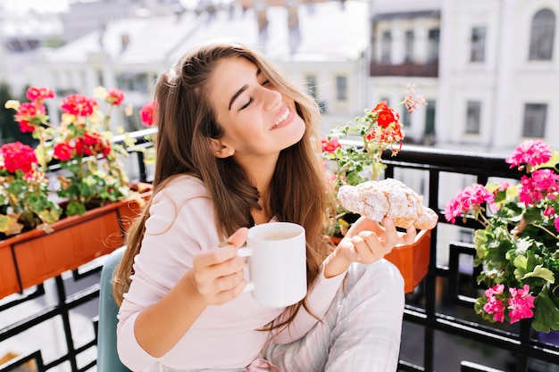 Retrato bastante joven con pelo largo desayunando en el balcón por la mañana. Ella sostiene una taza, un croissant, mantiene los ojos cerrados y se ve disfrutada.