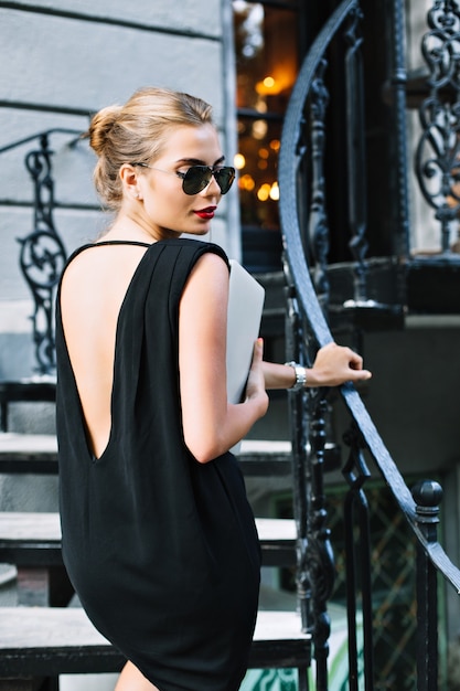 Foto gratuita retrato bastante modelo en vestido corto negro con la espalda desnuda en las escaleras al aire libre. ella está mirando hacia abajo.