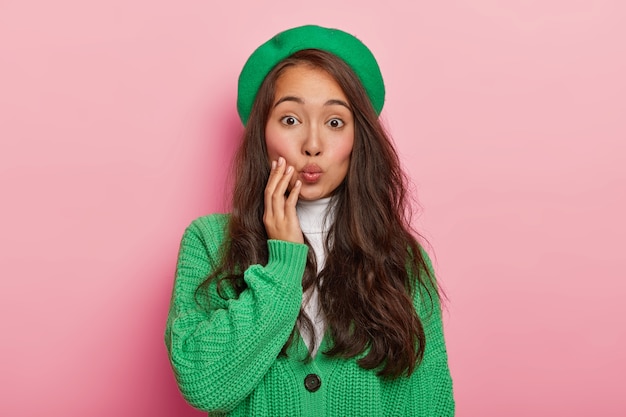 Retrato de bella dama asiática con cabello oscuro, mantiene los labios redondeados, quiere besar a su novio, viste un suéter y boina verde